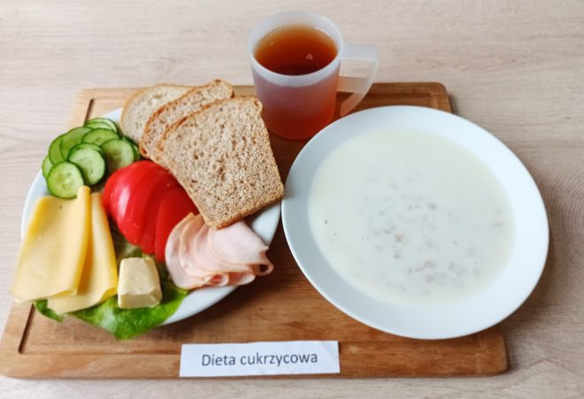 Na tacy znajduje się śniadanie w diecie cukrzycowej. Na talerzu ułożono kromki pełnoziarnistego chleba, plasterki żółtego sera, szynki drobiowej i masła, a także warzywa – pomidora, ogórek i liść sałaty. Obok znajduje się talerz z mlekiem i płatkami zbożowymi oraz kubek z herbatą. Przed tacą umieszczono napis „Dieta cukrzycowa”