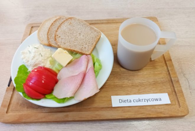 Śniadanie w diecie cukrzycowej: trzy kromki pieczywa jasnego i razowego, plasterki szynki, masło, twarożek z koperkiem, plaster pomidora i liście sałaty. Obok kubek kawy z mlekiem