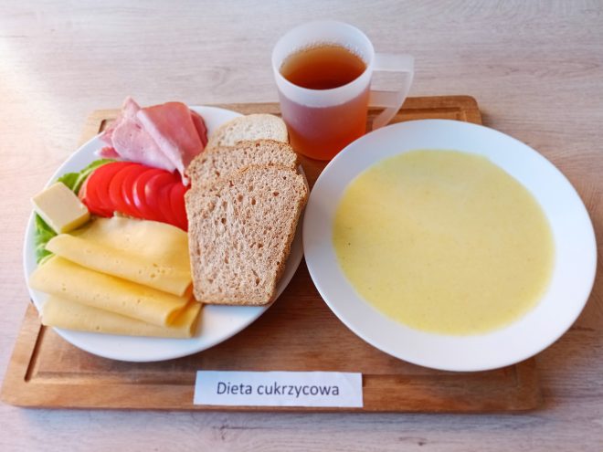 Śniadanie w diecie cukrzycowej. Na tacy talerz z ciemnym i jasnym pieczywem, plastrami sera, wędliną, pomidorem i sałatą, a także kawałkiem masła. Obok miseczka z zupą mleczną i kubek z herbatą