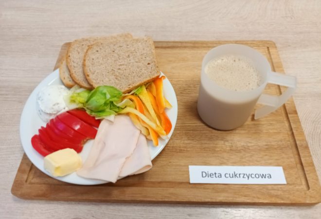 Na tacy z napisem „Dieta cukrzycowa” znajduje się talerz z pieczywem, plastrami pomidora, papryki i sałaty, plasterkami wędliny drobiowej, porcją masła i białym serkiem. Obok stoi kubek z kawą z mlekiem lub kawą zbożową