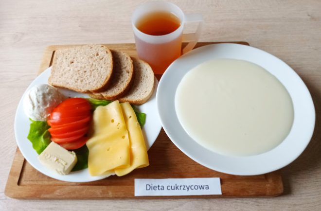 Na tacy z napisem „Dieta cukrzycowa” leży talerz z kilkoma kromkami pieczywa, plasterkami pomidora, żółtego sera i białego sera, porcją masła oraz listkiem sałaty. Obok znajduje się miska z białą kaszką mleczną i kubek z herbatą