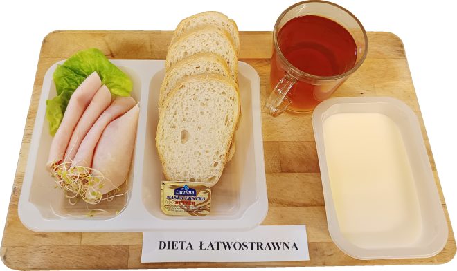 Na drewnianej tacy podano śniadanie w diecie łatwostrawnej. W przezroczystym pojemniku ułożono kilka plastrów chudej wędliny z dodatkiem sałaty i kiełków. Obok znajduje się porcją pieczywa pszennego, kostka masła oraz szklanka herbaty. Całość uzupełnia deser mleczny w prostokątnym pojemniku