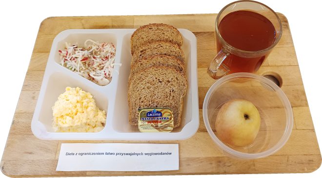 Na tacy leży pojemnik z trzema przegródkami: w jednej sałatka z tartej rzodkiewki, w drugiej pasta jajeczna, w trzeciej kilka kromek pełnoziarnistego chleba i porcja masła. Obok szklanka herbaty oraz plastikowy pojemnik z jabłkiem. Na kartce widnieje napis „Dieta z ograniczeniem łatwo przyswajalnych węglowodanów”
