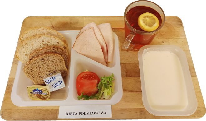 Na tacy ułożony zestaw śniadaniowy w ramach diety podstawowej. W zestawie znajdują się trzy kromki pieczywa pszennego i żytniego, porcja masła i sera topionego, kilka plastrów wędliny drobiowej, plaster pomidora z listkiem sałaty, kubek herbaty z plasterkiem cytryny oraz miseczka z białym napojem mlecznym lub zupą mleczną. Całość podana na drewnianej desce z podpisem „Dieta podstawowa”