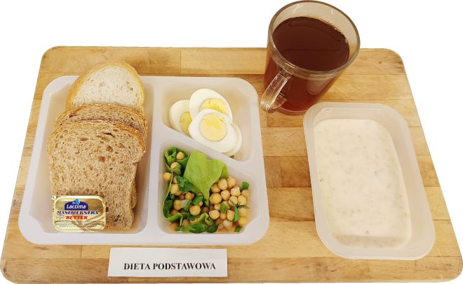 Na drewnianej tacy znajduje się śniadanie w pojemnikach dietetycznych opisane jako „Dieta podstawowa”. W zestawie widać kilka kromek pieczywa pełnoziarnistego, porcję masła, gotowane jajko pokrojone w plastry, sałatkę z ciecierzycą i zielonymi warzywami, kubek z herbatą oraz miseczkę z białą zupą mleczną lub owsianką. Zestaw jest przeznaczony dla pacjentów na standardowej diecie szpitalnej