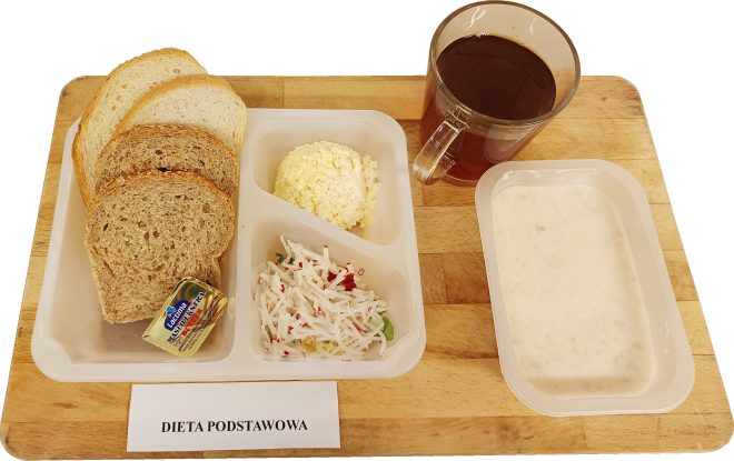Zestaw śniadaniowy w pojemnikach opisany jako „Dieta podstawowa”. Na tacy znajdują się kromki pieczywa jasnego i razowego, porcja masła, pasta jajeczna, surówka z białej rzodkwi i natki pietruszki, kubek herbaty oraz miseczka z białym jogurtem lub zupą mleczną. Całość ułożona na drewnianej desce