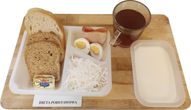 Zestaw śniadaniowy „Dieta podstawowa” podany na tacy. W skład wchodzą kromki pieczywa jasnego i ciemnego, porcja masła, gotowane jajko, plaster szynki, surówka z białej kapusty, napój w szklance oraz biały jogurt lub zupa mleczna w plastikowym pojemniku