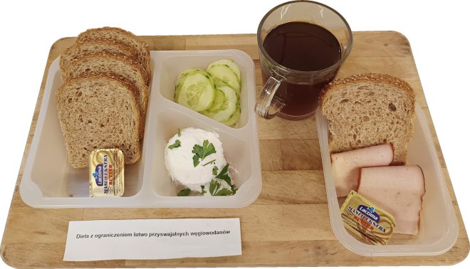 Na tacy oznaczonej napisem „Dieta z ograniczeniem łatwo przyswajalnych węglowodanów” podano zestaw śniadaniowy. W skład posiłku wchodzą kromki pieczywa pełnoziarnistego, plasterki ogórka, porcja twarogu udekorowanego natką pietruszki, kilka plastrów chudej wędliny oraz porcje masła. Do posiłku podano filiżankę kawy zbożowej lub herbaty. Całość ustawiona jest na drewnianej desce