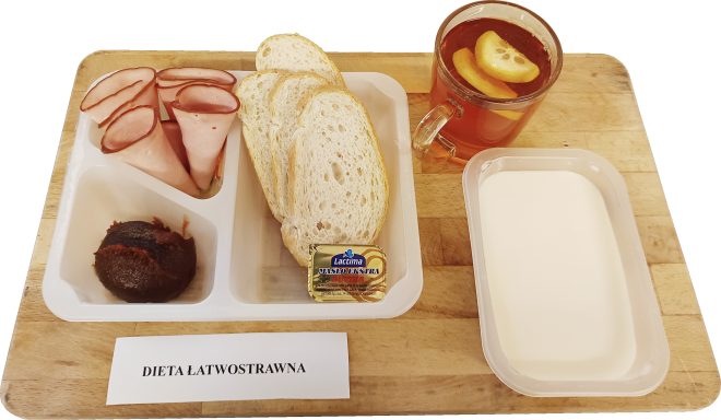 Zestaw śniadaniowy w plastikowej tacce: kilka plastrów szynki, porcja dżemu, kromki białego pieczywa, masło w jednorazowym opakowaniu. Obok przezroczysty kubek z herbatą i plasterkami cytryny oraz miseczka z białą zupą mleczną. Na desce pod spodem kartka z napisem „Dieta łatwostrawna”