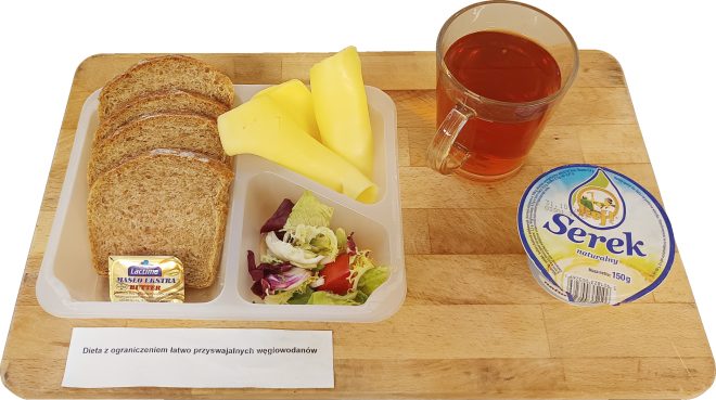 Zestaw śniadaniowy z czterema kromkami pełnoziarnistego chleba, masłem, dwoma plastrami sera żółtego, porcją sałatki z liści i warzyw, kubkiem herbaty oraz serkiem naturalnym w kubeczku. Na kartce przed tacą widnieje napis: „Dieta z ograniczeniem łatwo przyswajalnych węglowodanów”