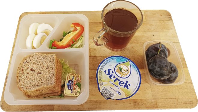 Śniadanie w diecie z ograniczeniem łatwo przyswajalnych węglowodanów. Na tacy znajdują się: kromki chleba pełnoziarnistego, masło, sałatka z warzyw, plasterki jajka na twardo, kawałki papryki i cebuli, serek naturalny w kubeczku, kubek z herbatą oraz kilka śliwek w pojemniku