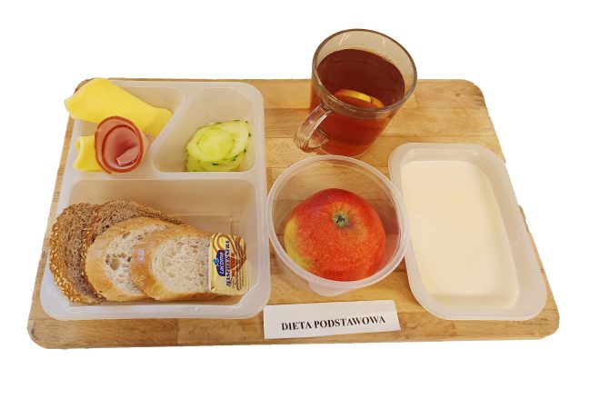 Na drewnianej desce znajduje się śniadanie opisane etykietą „Dieta podstawowa”. W zestawie podano kilka kromek jasnego i ciemnego pieczywa z porcją masła, plastry sera żółtego i szynki, kawałki świeżego ogórka oraz małą porcję ketchupu. Obok, w osobnych pojemnikach, znajduje się jabłko i miseczka z jogurtem lub kefirem. Do zestawu dołączono kubek z herbatą z cytryną