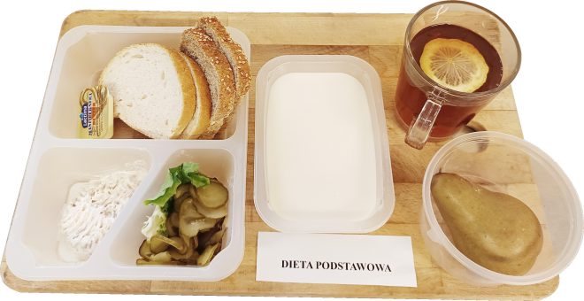 Na drewnianej tacy przygotowano śniadanie oznaczone jako „Dieta podstawowa”. W zestawie widać kromki chleba pszennego i żytniego z porcją masła, pastę rybną lub twarożek, sałatkę z ogórków kiszonych i liści sałaty, kubek herbaty z cytryną, pojemnik z gruszką oraz napój mleczny, prawdopodobnie kefir lub mleko. Posiłek stanowi przykład tradycyjnej diety szpitalnej