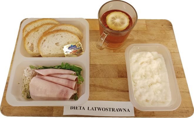 Na zdjęciu widać śniadanie w ramach diety łatwostrawnej: trzy kromki białego pieczywa z porcją masła, wędliną drobiową podaną z sałatą i kiełkami, a także pojemnik z białym twarożkiem. Do picia herbata z plastrem cytryny, całość ustawiona na drewnianej tacy