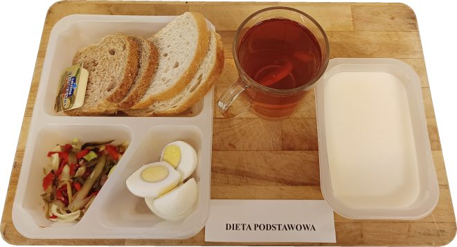 Na tacy znajduje się przykładowe śniadanie w diecie podstawowej. Zestaw obejmuje kilka kromek pieczywa jasnego i ciemnego, porcję masła, gotowane jajko, surówkę z warzyw (m.in. papryki, ogórka i kapusty) oraz napój – herbatę owocową. W oddzielnym pojemniku znajduje się jogurt naturalny lub mleko. To pełnowartościowy, urozmaicony posiłek zapewniający energię na początek dnia
