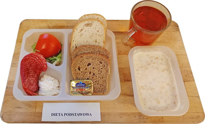 Na tacy ułożony jest zestaw śniadaniowy w diecie podstawowej. W plastikowym pojemniku znajdują się plasterki salami, twarożek, kawałek pomidora z listkiem sałaty, trzy kromki pieczywa jasnego i ciemnego oraz porcja masła. Obok pojemnika stoi kubek z herbatą i miseczka z mlekiem i płatkami owsianymi. Przed tacą widoczny jest napis „Dieta podstawowa”