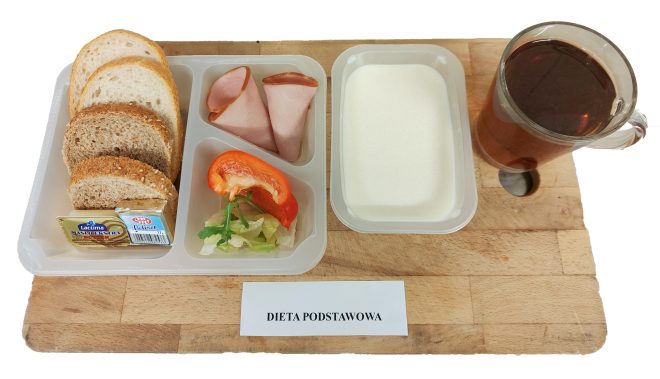Taca z posiłkiem w diecie podstawowej. W zestawie kilka kromek chleba jasnego i pełnoziarnistego, masło, serek topiony, dwa plasterki szynki, sałatka z warzyw oraz kubek herbaty i porcja jogurtu naturalnego