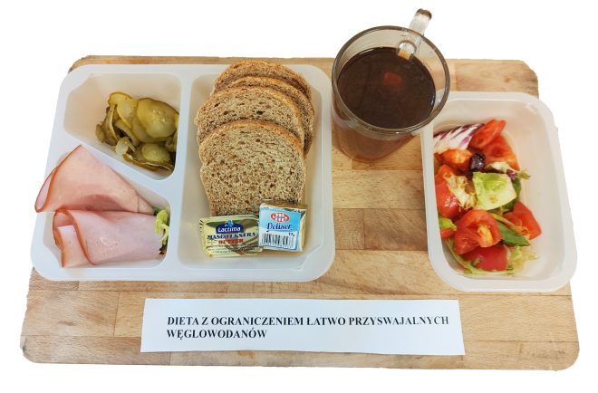 Zestaw śniadaniowy w diecie z ograniczeniem łatwo przyswajalnych węglowodanów. Na tacy znajdują się trzy kromki chleba razowego, dwa plasterki wędliny, porcje masła i serka topionego, ogórki kiszone oraz sałatka z pomidorów i sałaty. Obok stoi szklanka z herbatą. Przed tacą leży kartka z napisem „Dieta z ograniczeniem łatwo przyswajalnych węglowodanów”