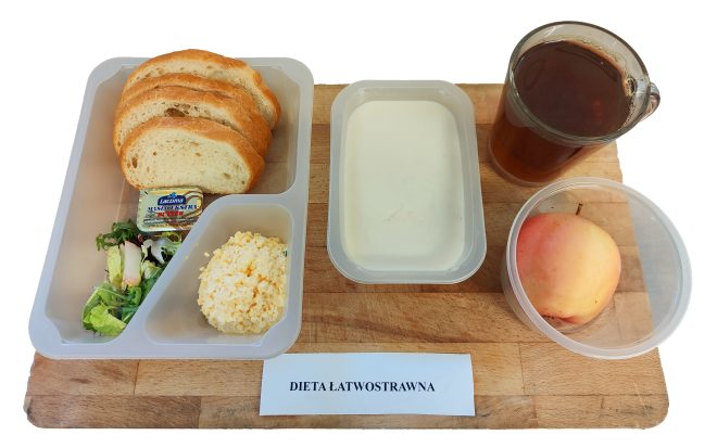 Na tacy z napisem „DIETA ŁATWOSTRAWNA” znajduje się plastikowy zestaw z kilkoma kromkami białego pieczywa, porcją sałatki, masłem i pastą jajeczną. Obok leży jabłko w pojemniku, kubek z herbatą i pojemnik z jasną zupą mleczną