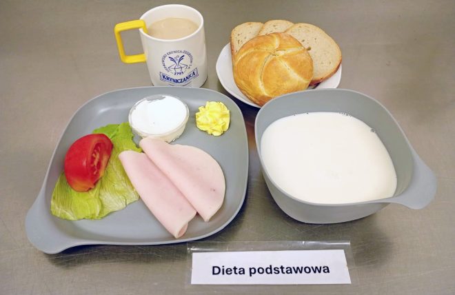 Śniadanie: dwa plasterki wędliny drobiowej na liściu sałaty z kawałkiem pomidora, porcja serka i masła. Obok miska z mlekiem, bułka i kromki chleba pszennego oraz kubek kawy z mlekiem. Na stoliku kartka z napisem Dieta podstawowa