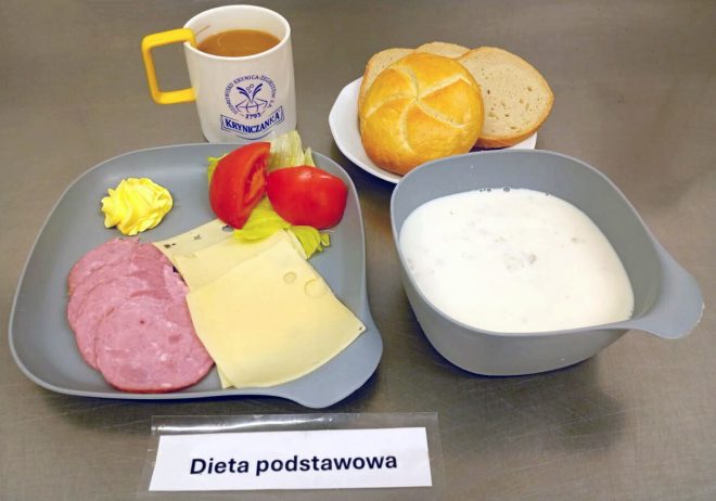 Na zdjęciu przedstawiono śniadanie w ramach „Diety podstawowej”. Na szarym talerzu leżą plasterki wędliny, dwa plasterki żółtego sera, masło, liść sałaty i kawałki pomidora. Obok znajduje się miska z mleczną zupą lub płatkami, talerzyk z bułką i kromkami pieczywa oraz kubek z kawą z mlekiem w białym naczyniu z żółtym uchem i logo „Kryniczanka”. Zestaw znajduje się na metalicznym blacie