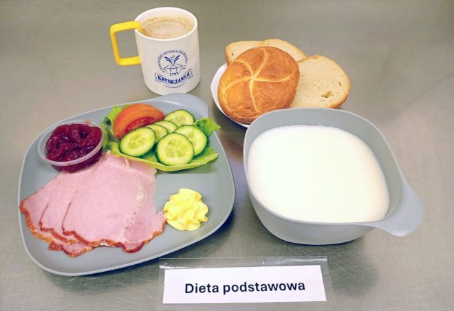 Na zdjęciu widoczny jest zestaw śniadaniowy w ramach diety podstawowej. Na szarym talerzu ułożono kilka plasterków wędliny, porcję masła, liść sałaty z plasterkami świeżego ogórka i kawałkiem pomidora oraz mały pojemnik z porcją czerwonej konfitury. Obok znajduje się miska z mlekiem, biały talerzyk z bułką i dwiema kromkami chleba pszennego oraz kubek z kawą z mlekiem w naczyniu z logo „Kryniczanka”. Przed posiłkiem umieszczono kartkę z napisem „Dieta podstawowa”