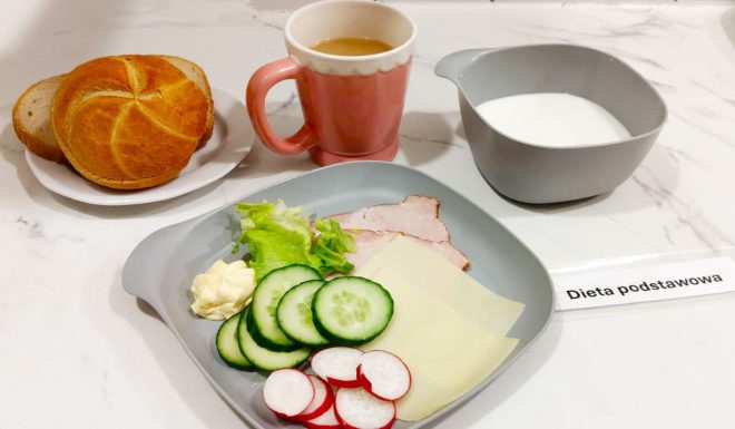 Na talerzu leży bułka kajzerka i kilka kromek chleba pszennego. Obok, w różowym kubku, znajduje się kawa z mlekiem, a w szarej miseczce porcja mleka lub zupy mlecznej. Na talerzu z szarego tworzywa ułożono plasterki ogórka i rzodkiewki, dwa plastry żółtego sera, szynkę, liść sałaty i porcję masła. Przed zestawem leży karteczka z napisem „Dieta podstawowa”