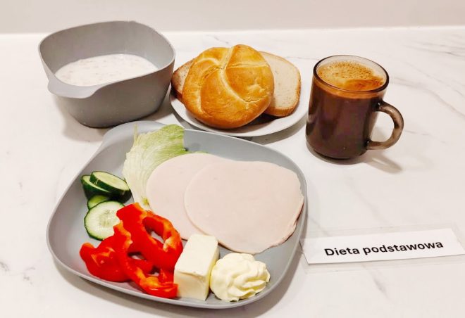 Na szarej miseczce ułożono dwa plastry szynki drobiowej, kilka kawałków czerwonej papryki, plasterki ogórka, liść sałaty, kostkę sera i porcję majonezu. W tle widać talerzyk z bułką kajzerką i kromkami chleba oraz kubek z kawą z pianką. Obok znajduje się miseczka z płatkami owsianymi na mleku. Zestaw opatrzono karteczką z napisem „Dieta podstawowa”