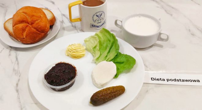 Zestaw śniadaniowy oznaczony jako „Dieta podstawowa”. Na talerzu bułka i kromki chleba, sałata, plaster sera, porcja masła, pasty warzywnej i ogórek kiszony. Obok filiżanka z mlekiem i kubek z gorącym napojem, prawdopodobnie kawą zbożową