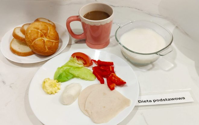 Śniadanie na białym talerzu: plasterki wędliny drobiowej, masło, biały serek, sałata, pomidor i papryka czerwona. Obok pieczywo – bułka i kromki chleba, kubek z kawą z mlekiem oraz miseczka z zupą mleczną. Na stole kartka z napisem „Dieta podstawowa”