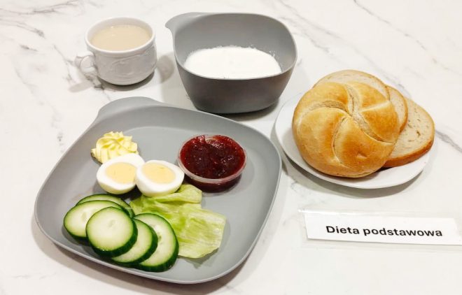Na szarym talerzu jajko na twardo, plasterki ogórka, liść sałaty, porcja masła i dżem w małym pojemniczku. Obok bułka i kromki chleba, miseczka z białą zupą mleczną oraz filiżanka z kawą z mlekiem. Na stole widoczna kartka z napisem „Dieta podstawowa”