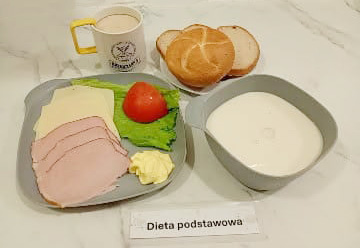 Śniadanie składające się z kromek chleba i bułki, sałaty z plastrem pomidora, plastrów wędliny drobiowej i sera żółtego, porcji masła oraz miseczki z zupą mleczną. Do posiłku podano kubek z kawą z mlekiem. Na kartce przed zestawem znajduje się napis „Dieta podstawowa”