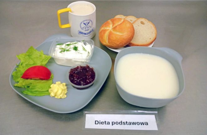 Śniadanie w diecie podstawowej. Na tacy znajduje się talerz z sałatą, plasterkiem pomidora i porcją masła, twarożkiem z koperkiem oraz dżemem. Obok stoją bułka, kromki chleba, kubek z napojem mlecznym oraz miseczka z zupą mleczną. Przed tacą umieszczono kartkę z napisem „Dieta podstawowa”