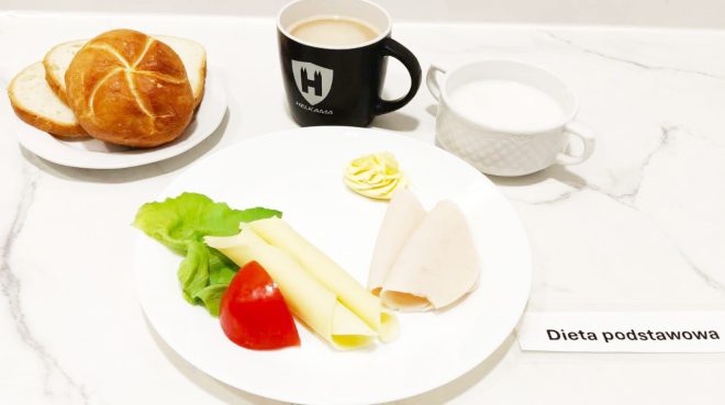 Śniadanie w ramach diety podstawowej: na białym talerzu znajdują się dwa plastry wędliny drobiowej, dwa zrolowane plastry sera żółtego, liść sałaty i kawałek pomidora, a także porcja masła. Obok ustawiono bułkę kajzerkę i dwie kromki jasnego chleba, czarny kubek z kawą z mlekiem oraz filiżankę z białym napojem mlecznym. Na stole widoczna jest karteczka z napisem „Dieta podstawowa”
