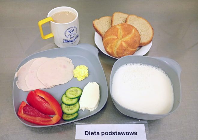 Na zdjęciu widać śniadanie w ramach diety podstawowej. Na tacy znajdują się trzy plasterki drobiowej wędliny, porcja masła, biały ser, kilka plasterków świeżego ogórka i kawałki czerwonej papryki. Do zestawu dołączono miseczkę z białym napojem mlecznym, bułkę kajzerkę oraz trzy kromki chleba pszennego. Obok stoi kubek z kawą z mlekiem w żółto-białym kubku z logo „Kryniczanka” i kartka z napisem „Dieta podstawowa”
