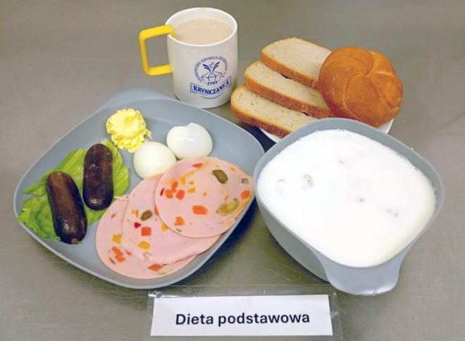 Śniadanie w ramach diety podstawowej: na szarej tacy ułożono dwa plastry wędliny z dodatkiem warzyw, dwa parówki, dwa jajka na twardo, liść sałaty oraz porcję masła. Obok znajduje się miseczka z mlekiem, kubek z kawą z mlekiem, a na talerzyku – bułka kajzerka i kromki jasnego pieczywa. Całość oznaczona jest karteczką z napisem „Dieta podstawowa”