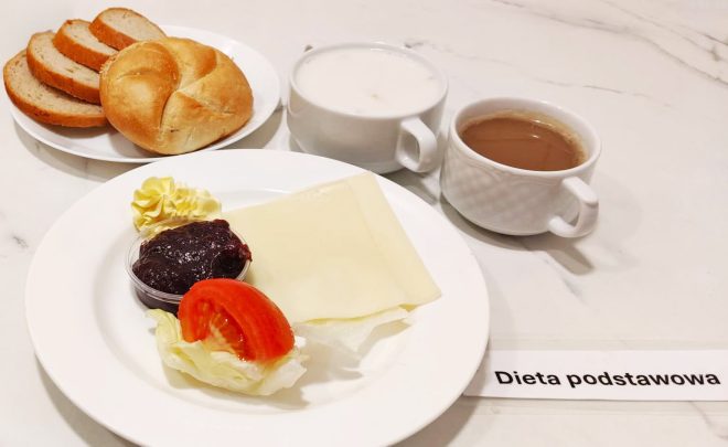 Na białym talerzu znajduje się plaster żółtego sera, porcja masła, liść sałaty z kawałkiem pomidora oraz mały kubeczek konfitury z czarnej porzeczki. Obok talerzyk z kajzerką i trzema kromkami chleba, filiżanka mleka i filiżanka kawy z mlekiem. Na blacie leży karteczka z napisem „Dieta podstawowa”