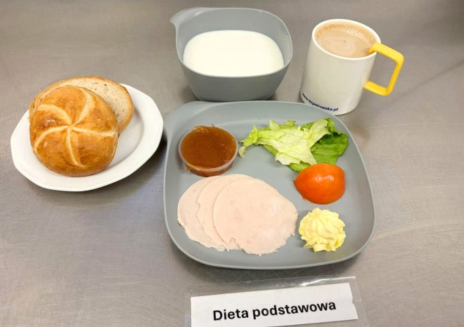 Na szarym talerzu znajdują się trzy cienkie plastry wędliny drobiowej, porcje masła i sałaty lodowej, połówka pomidora oraz mały kubeczek dżemu jabłkowego. Obok na osobnym talerzyku leży kajzerka i dwa kromki chleba pszenno-żytniego. W szarym kubku mleko, a w białym kubku z żółtym uchem – kawa z mlekiem. Na stoliku leży karteczka z napisem „Dieta podstawowa”