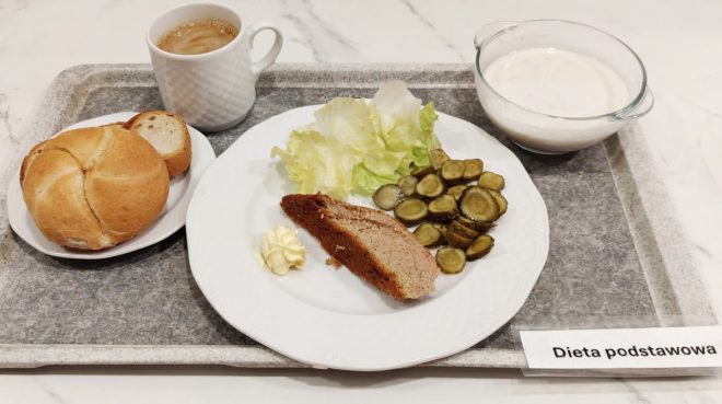 Na tacy znajduje się śniadanie: bułka i kilka kromek chleba, kawa z mlekiem w białym kubku, porcja pieczeni z masłem, sałata i plasterki ogórków kiszonych. W przezroczystej miseczce widoczna biała zupa mleczna lub sos. Po prawej stronie kartka z napisem „Dieta podstawowa”