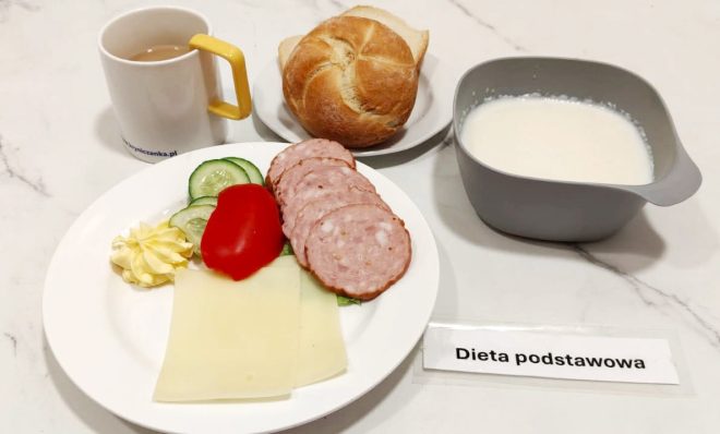 Na talerzu ułożono plasterki kiełbasy, żółty ser, kawałek pomidora, plasterki ogórka oraz rozetkę masła. Do zestawu podano kajzerkę, kubek kawy z mlekiem oraz szarą miseczkę z mlecznym napojem. Całość oznaczona jako posiłek w diecie podstawowej