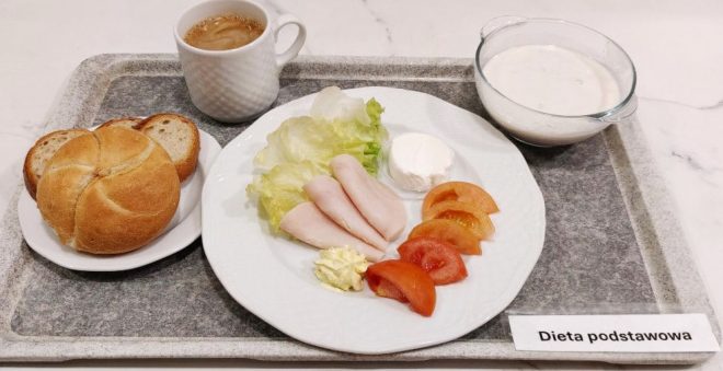 Na tacy z napisem „Dieta podstawowa” znajduje się talerz z liśćmi sałaty, kawałkami czerwonej papryki, porcją twarożku ziarnistego i masła. Obok stoją miseczka z zupą mleczną z kaszą manną, filiżanka kawy z mlekiem oraz pieczywo – kilka kromek chleba i bułka kajzerka