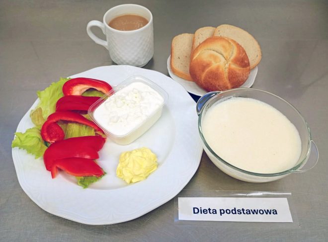Na tacy z napisem „Dieta podstawowa” znajduje się talerz z liśćmi sałaty, kawałkami czerwonej papryki, porcją twarożku i masła. Obok ustawiono miseczkę z zupą mleczną z kaszą manną, filiżankę kawy z mlekiem oraz pieczywo: bułkę i kilka kromek chleba