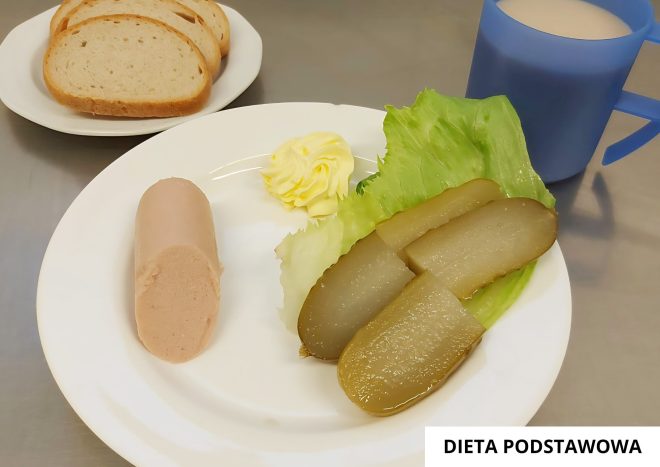 Porcja śniadaniowa: kawałek pasztetu drobiowego, trzy plastry ogórka kiszonego na liściu sałaty, porcja masła. Obok talerzyk z trzema kromkami chleba pszennego oraz kubek z kawą z mlekiem. Na stole kartka z napisem Dieta podstawowa