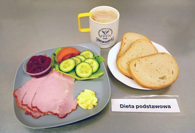 Na zdjęciu widoczny jest zestaw śniadaniowy w ramach diety podstawowej. Na szarym talerzu ułożono kilka plasterków wędliny, porcję masła, kilka plasterków świeżego ogórka i kawałek pomidora na liściu sałaty oraz mały pojemniczek z porcją czerwonej konfitury. Obok, na białym talerzyku, znajdują się trzy kromki pieczywa pszennego. W tle stoi kubek z napojem mlecznym lub kawą z mlekiem w filiżance z logo „Kryniczanka”. Na stole leży karteczka z napisem „Dieta podstawowa”
