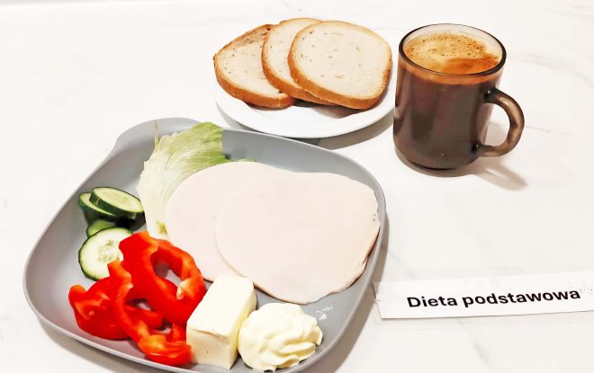 Zestaw śniadaniowy opisany jako „Dieta podstawowa”. Na talerzu leżą plastry wędliny drobiowej, masło, majonez, liść sałaty, plasterki ogórka i papryki. Obok talerza znajdują się kromki chleba jasnego na osobnym talerzyku oraz kubek z kawą z mlekiem. Całość ustawiona na jasnym blacie