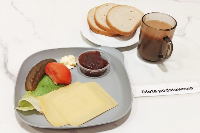 Zestaw kolacyjny opisany jako „Dieta podstawowa”. Na talerzu znajdują się dwa plasterki sera żółtego, sałata, plaster pomidora, ogórek kiszony, porcja masła i mały pojemniczek z dżemem. Obok ułożone są kromki pieczywa jasnego na osobnym talerzyku oraz kubek z kawą z mlekiem. Całość na jasnym blacie