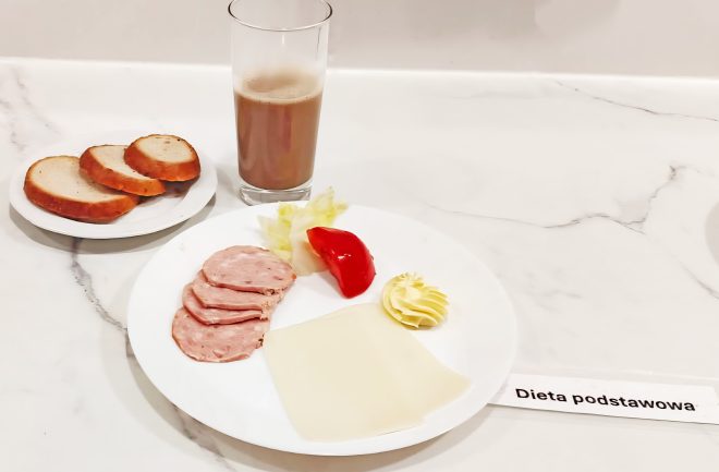 Na białym blacie stoi talerz z posiłkiem oznaczonym etykietą „Dieta podstawowa”. Na talerzu znajdują się trzy plasterki wędliny, dwa plasterki sera żółtego, kawałek czerwonej papryki, odrobina sałaty i porcja masła. Obok, na małym talerzyku, leżą trzy kromki jasnego pieczywa. W tle stoi szklanka z kakao lub kawą z mlekiem. Całość ułożona jest schludnie, w szpitalnym lub dietetycznym kontekście żywieniowym