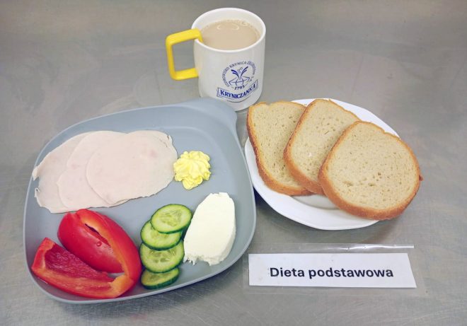 Na zdjęciu przedstawiono śniadanie w ramach diety podstawowej. Na szarej tacy ułożono plasterki drobiowej wędliny, porcję masła, kawałek białego sera, kilka plasterków świeżego ogórka i kawałki czerwonej papryki. Obok znajdują się trzy kromki białego pieczywa oraz kubek z kawą z mlekiem w żółto-białym kubku z logo „Kryniczanka”. Na stole widoczna jest również karteczka z napisem „Dieta podstawowa”