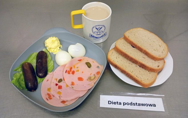 Śniadanie w ramach diety podstawowej: na talerzu trzy kromki chleba pszennego, obok talerz z dwiema jajkami na twardo, plastrami mortadeli z warzywami, dwiema kiszonymi ogórkami, porcją masła i listkiem sałaty. Do picia kawa z mlekiem w kubku z napisem „Kryniczanka”