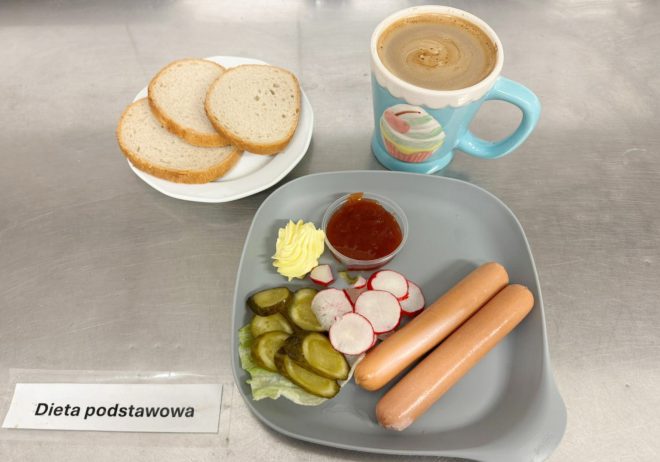 Na zdjęciu widoczny jest zestaw posiłku oznaczony jako „Dieta podstawowa”. Na szarym talerzu znajdują się dwa parówki, kilka plasterków rzodkiewki, ogórek kiszony w plastrach, listek sałaty, porcja masła oraz mały pojemniczek z ketchupem. Obok talerza stoi filiżanka z napojem – prawdopodobnie kawa z mlekiem – w błękitnym kubku z motywem babeczki. Z lewej strony na małym białym talerzyku leżą trzy kromki białego pieczywa. Wszystko ustawione jest na metalowym blacie